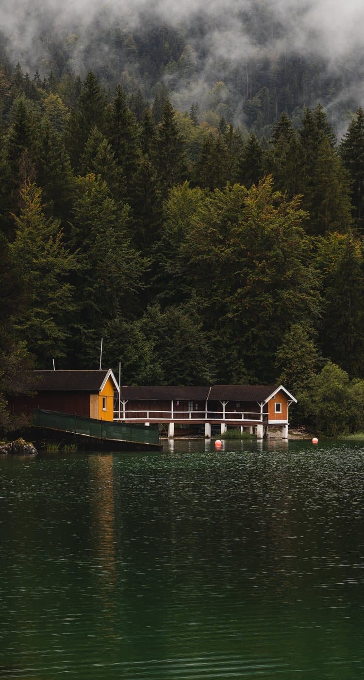 A timber boathouse on the shoreline of a still alpine lake, dark evergreen forest and low cloud behind it