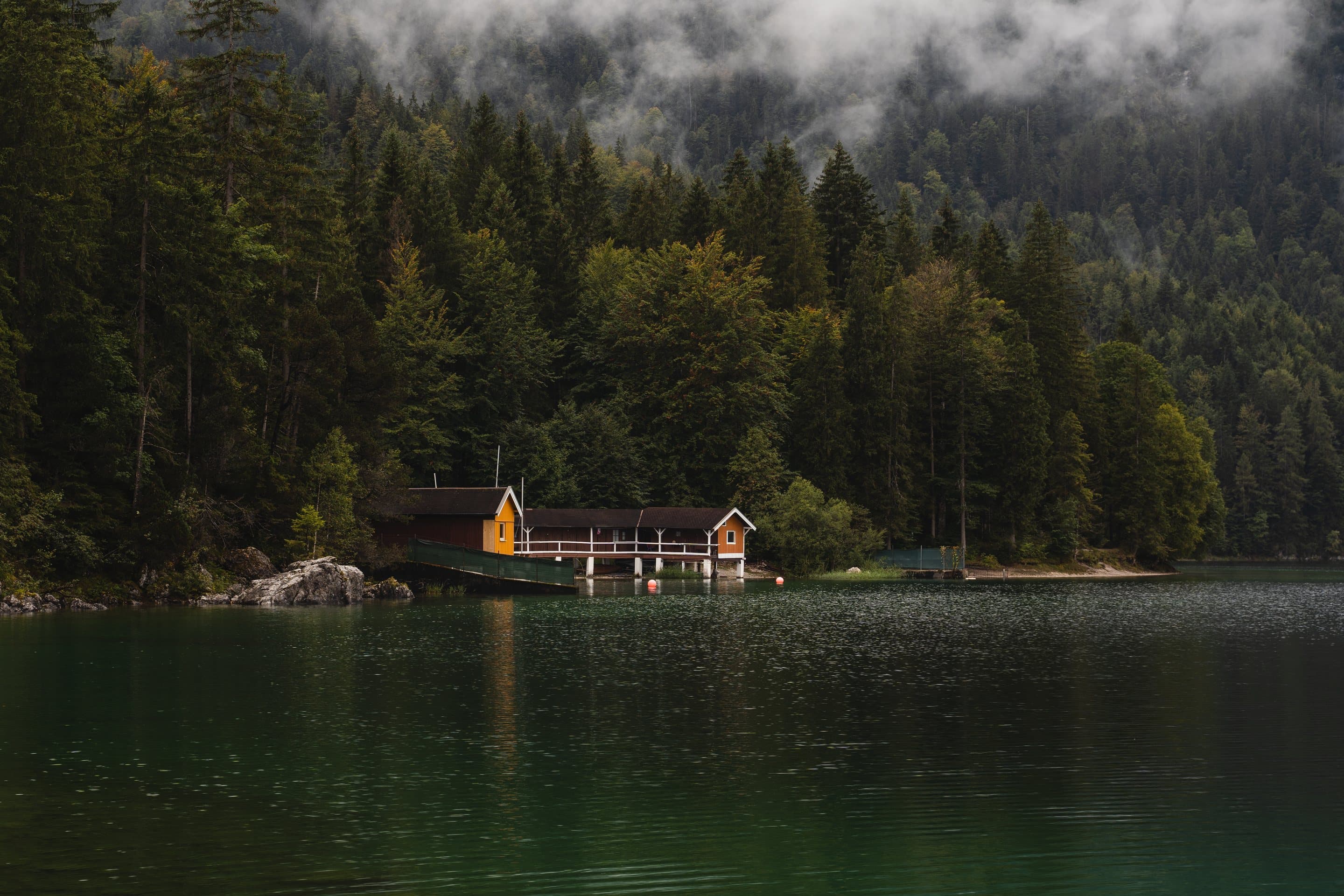 A timber boathouse on the shoreline of a still alpine lake, dark evergreen forest and low cloud behind it